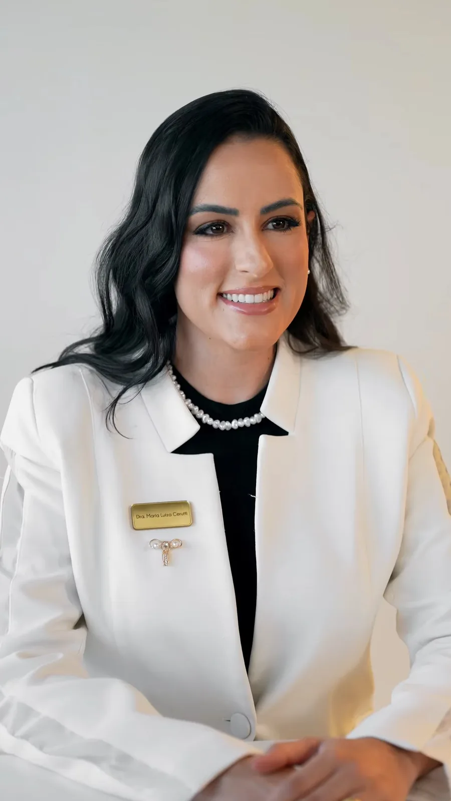Dra. Mallu Cerutti sorrindo, sentada, de blazer branco com plaqueta de identificação, colar de pérolas e um broche em forma de útero