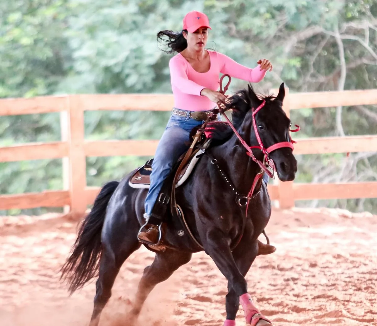 Dra. Mallu Cerutti a galope num cavalo preto, de boné, camisa e caneleiras rosa, numa pista de areia